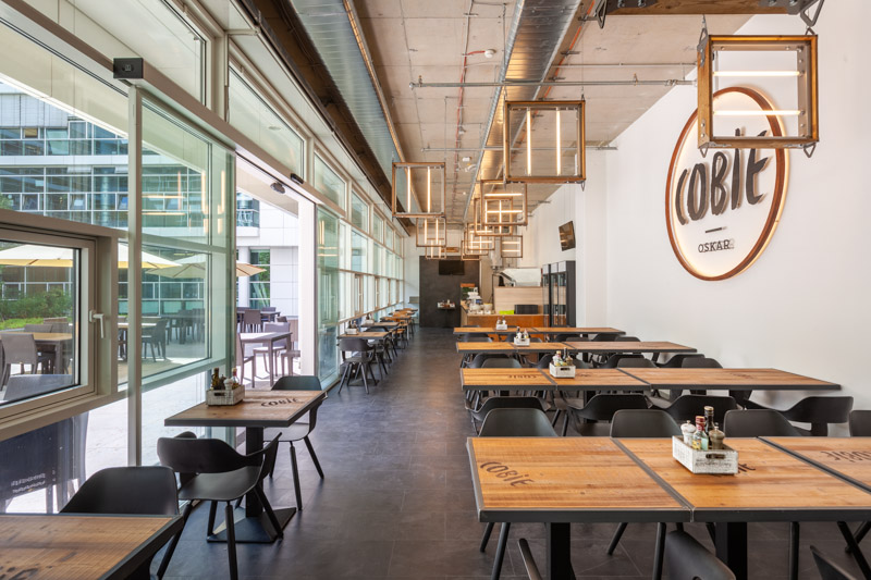 Interior of the Cobie restaurant in the OSKAR quarter Munich, long dining space in industrial style with wooden tables, black chairs, distinctive pendant lights and a large window front to the terrace, captured by Max Sydow