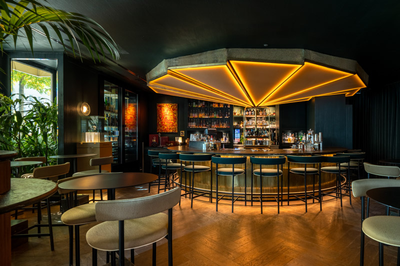 Interior of the ORY Bar at the Mandarin Oriental Munich, elegant oval bar counter with petrol-blue bar stools beneath a golden fan-shaped ceiling light, dark green walls, backlit whisky and spirits shelves and lush green plants, captured by Max Sydow