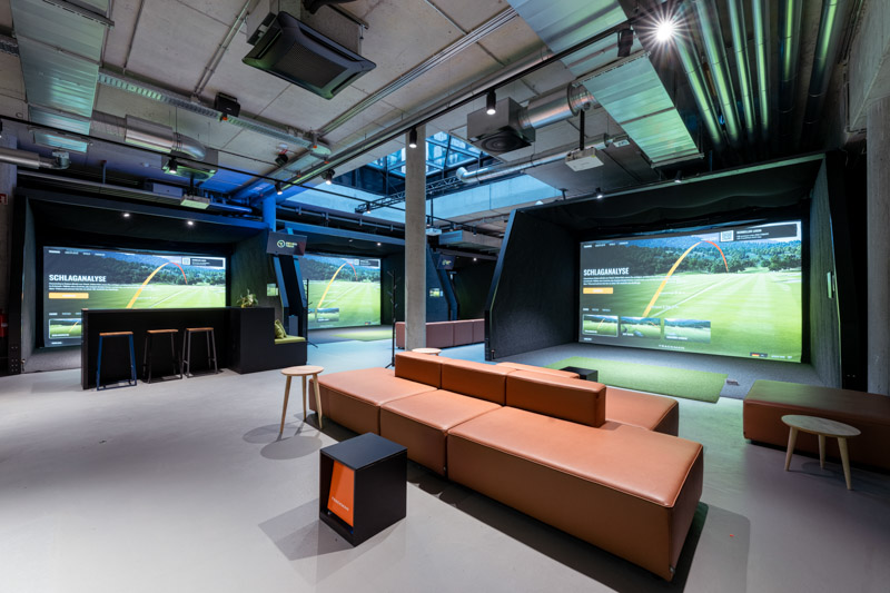Interior of the Seven Tees Indoor Golf & Bar Munich, modern golf simulator lounge in industrial style with large TrackMan projection screens, brown leather couch and concrete ceiling, captured by Max Sydow