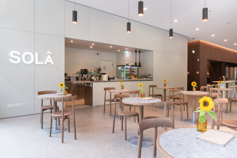 Interior of the SOLÂ Café in Munich, bright modern café with round terrazzo tables, wooden chairs and sunflowers, illuminated SOLÂ logo on the wall, captured by Max Sydow