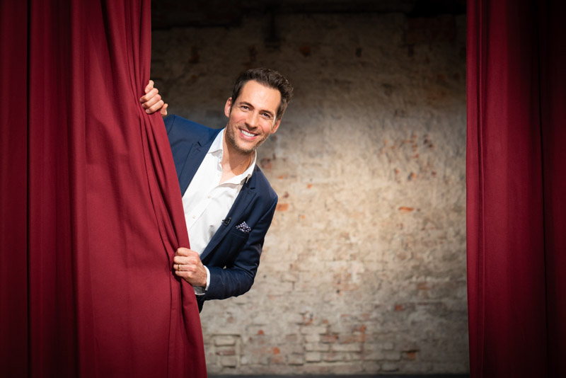 Editorial portrait of TV presenter Alexander Mazza in a blue suit, smiling as he peeks out from behind a red theatre curtain, captured by Max Sydow