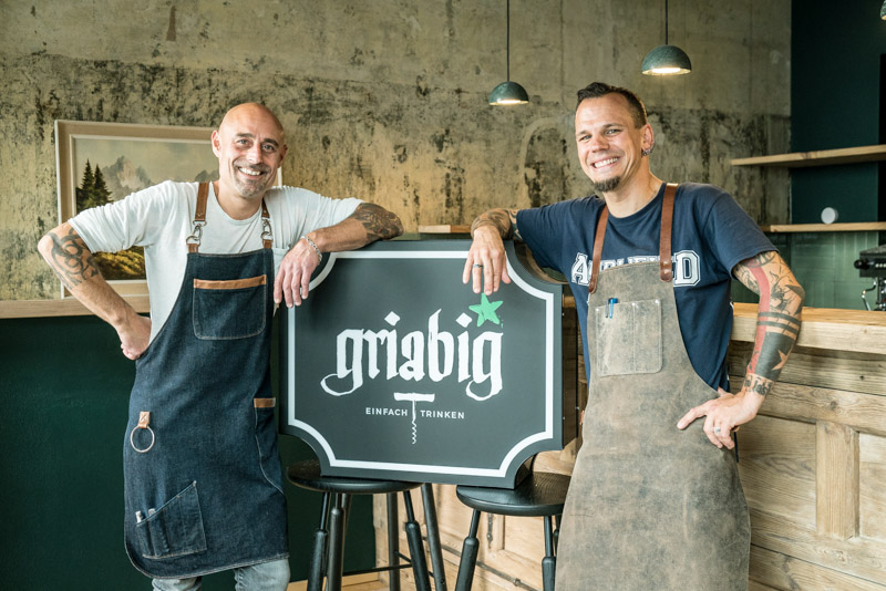Portrait of restaurateurs Bernd Arold and Holger Baier with aprons and tattoos next to the Griabig logo in their Munich wine bar, captured by Max Sydow